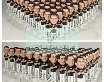 May include: A large group of custom bobbleheads, all wearing black shirts with their arms crossed, and white pants. The bobbleheads are arranged in a heart shape on a white surface.