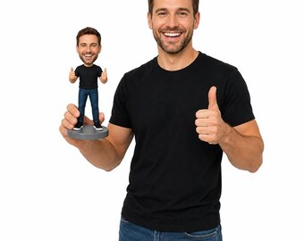 May include: A miniature figurine of a man in a black t-shirt and blue jeans giving a thumbs-up, held by a man in a black t-shirt and blue jeans. The figurine is on a gray base. Both men are smiling.
