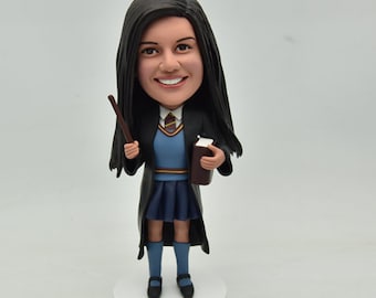 May include: A custom figurine of a person dressed in a black robe, blue shirt, and blue skirt. The figurine is holding a brown book and a brown wand. The figurine has long black hair and is smiling.