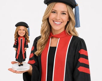 May include: A woman in a graduation gown and cap holds a bobblehead of herself. The gown is black with red accents and a white stripe. The bobblehead wears a matching gown and cap. The cap is black with a blue tassel.
