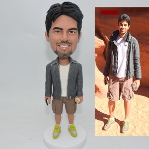 May include: A custom bobblehead figurine of a person wearing a grey jacket, brown shorts, and green trainers. The figurine is holding a phone in one hand and a bottle of water in the other. The figurine is standing on a white base.