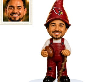 May include: A custom bobblehead figurine featuring a person with a beard wearing a white long-sleeve shirt and maroon overalls. The figure wears a tall, pointed maroon hat with an Arizona Cardinals logo and holds a brown football in one hand.