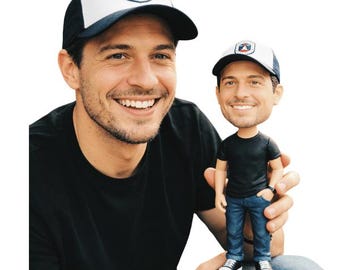 May include: A person wearing a black t-shirt and a white and navy blue baseball cap with a logo. The person is holding a bobblehead doll wearing a black t-shirt, blue jeans, and a matching cap.