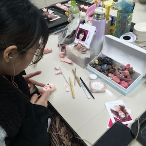 May include: A person working on a doll's face, with tools and materials on a white table. The table includes a box of colorful beads, a tray of doll heads, and a photo of a woman. A smartphone is also on the table.