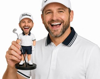 May include: A man wearing a white polo shirt and a white Titleist hat smiles broadly, holding a bobblehead of a golfer. The bobblehead wears a similar outfit and hat, holding a golf club. The bobblehead is on a black base.