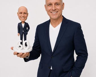 May include: A man in a navy blue suit smiles, holding a bobblehead figurine of himself. The figurine, wearing a similar suit, stands on a white base with the number "60". The background is plain white.
