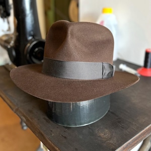 Könnte beinhalten: Ein brauner Fedora-Hut mit schwarzem Band, der auf einer dunklen Oberfläche ruht. Der Hut hat eine breite Krempe und ein klassisches, zeitloses Design. Der Hut ist wahrscheinlich aus Filz oder einem ähnlichen Material.