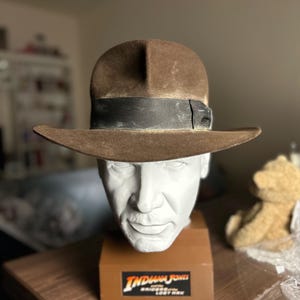 Könnte beinhalten: Ein brauner Filzhut mit schwarzem Band, getragen von einer weißen Gipsbüste von Indiana Jones. Die Büste steht auf einem braunen Sockel mit dem aufgedruckten Text "Indiana Jones Jäger des verlorenen Schatzes".