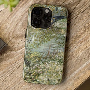 Peut inclure: Coque de smartphone avec une peinture de paysage aux tons verts, blancs et bleus. La coque a une finition brillante et repose sur une surface en bois. Un appareil photo noir et une plante verte sont &eacute;galement visibles.