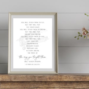 May include: Framed print with a light gray border, featuring a quote by St. Teresa of Calcutta. The text is in black, with a vintage airplane illustration. The print is displayed on a wooden surface with flowers in the background.