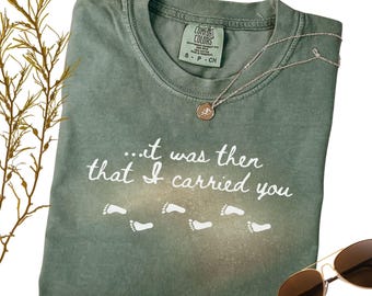 Puede incluir: Una camiseta Comfort Colors verde musgo con el texto "...it was then that I carried you" y huellas blancas. Una cadena de plata con un colgante y gafas de sol también están en la imagen.