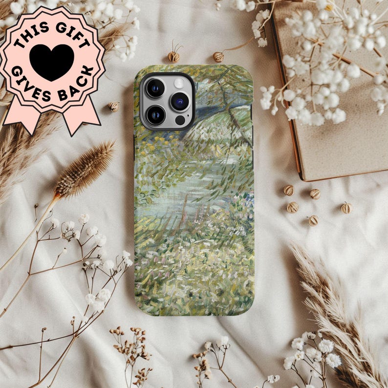 Peut inclure: Une coque de t&eacute;l&eacute;phone avec une peinture de paysage dans des tons de vert, blanc et bleu. L'&eacute;tui a une d&eacute;coupe noire pour l'objectif de l'appareil photo et un badge rose avec le texte "THIS GIFT GIVES BACK". L'&eacute;tui est entour&eacute; de fleurs s&eacute;ch&eacute;es et d'un livre.