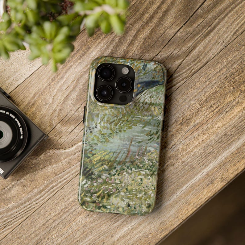 Peut inclure: Coque de smartphone avec une peinture de paysage aux tons verts, blancs et bleus. La coque a une finition brillante et repose sur une surface en bois. Un appareil photo noir et une plante verte sont &eacute;galement visibles.