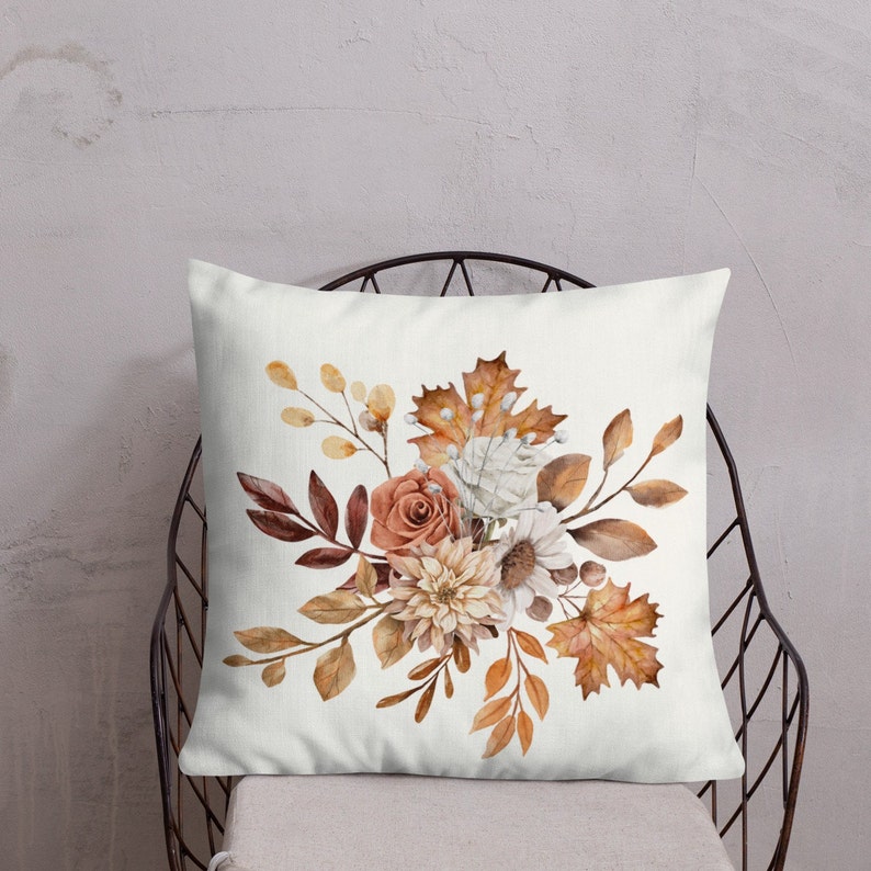 Puede incluir: Una almohada blanca con un dise&ntilde;o floral de acuarela que presenta un ramo de flores naranjas, blancas y marrones con hojas verdes. Las flores est&aacute;n dispuestas en un racimo y est&aacute;n rodeadas de hojas.