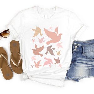 May include: White t-shirt with a pattern of pink, brown, and peach doves. The doves are all facing the same direction and are arranged in a random pattern.