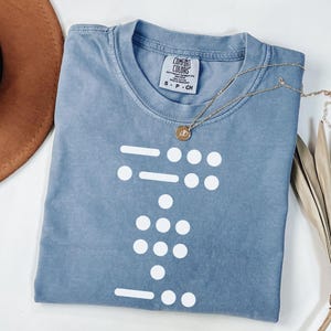 May include: A light blue t-shirt featuring a white graphic of a braille pattern. The shirt has a tag that reads "Comfort Colors" and "S - P - CH".