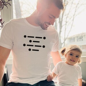 May include: A white t-shirt featuring a black design of a series of dots and dashes, resembling Morse code. A child is wearing a white t-shirt and holding the hand of the person wearing the t-shirt with the design.
