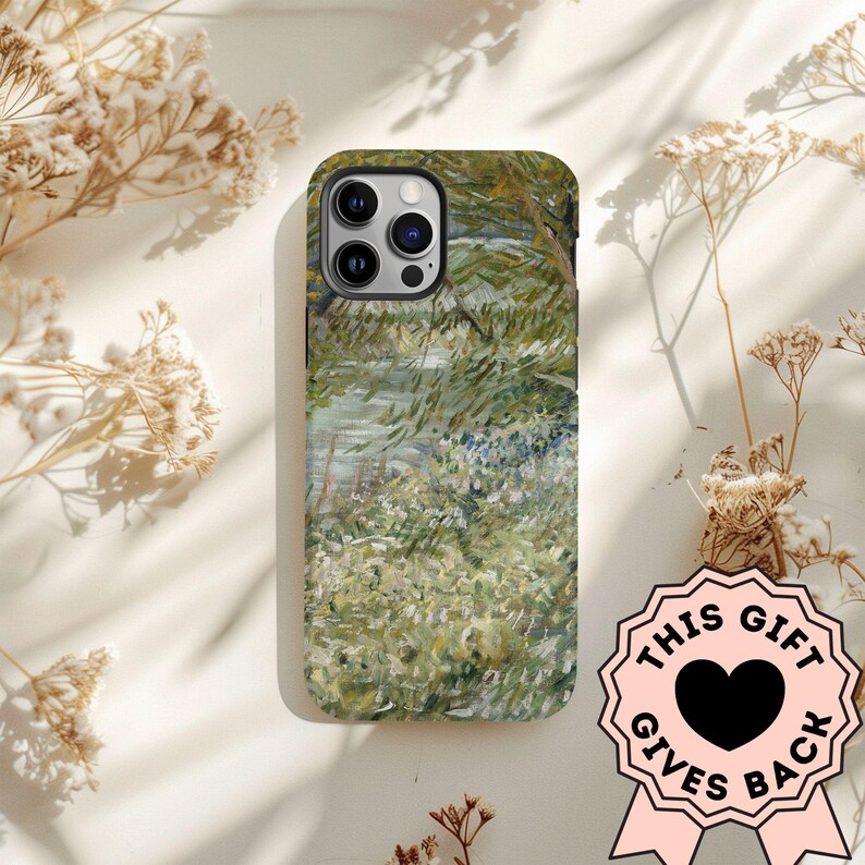 Peut inclure: Coque de t&eacute;l&eacute;phone avec une peinture de paysage dans les tons de vert, blanc et bleu. L'&eacute;tui a un module cam&eacute;ra noir et un ruban rose avec le texte "THIS GIFT GIVES BACK" et un c&oelig;ur noir.