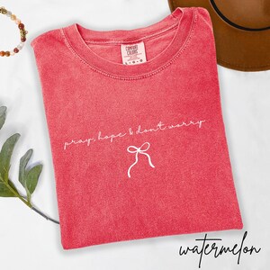 May include: A red t-shirt with the words "pray, hope & don't worry" and a bow graphic in white. The shirt is labeled "Comfort Colors" and the word "watermelon" is written in cursive at the bottom.