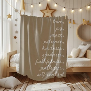 May include: A beige throw blanket with white script that reads "love, joy, peace, patience, kindness, goodness, faithfulness, gentleness, self-control." The blanket is hanging from a clothesline in a child's bedroom.