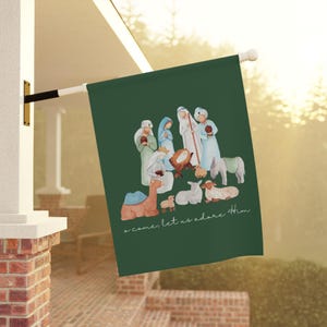 May include: A green outdoor flag featuring a watercolor illustration of the Nativity scene. The image includes figures of Mary, Joseph, and baby Jesus, along with animals. The text "o come, let us adore Him" is written in a script font.