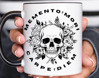 May include: White ceramic mug with a black handle and rim, featuring a skull surrounded by flowers and the phrases "Memento Mori" and "Carpe Diem" in black lettering. The mug is held in a person's hands.