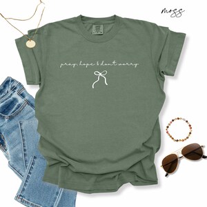 May include: An olive green t-shirt with the words "pray, hope & don't worry" in a delicate script, along with a bow graphic. The shirt is styled with blue jeans, sunglasses, a necklace, and a bracelet.