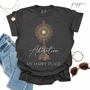 May include: A charcoal gray short-sleeve t-shirt featuring a gold-toned graphic of a monstrance. The word Adoration is printed in white script above the text MY HAPPY PLACE in white capital letters. The shirt is labeled as pepper color.