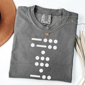 May include: A grey t-shirt with a white design of a braille pattern. The shirt has a tag that says "Comfort Colors" and "S - P - CH".