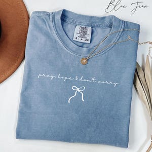 May include: A blue jean-colored t-shirt with the words "pray, hope & don't worry" in white script, along with a bow graphic. A gold necklace with a pendant is draped across the shirt. The shirt is a Comfort Colors brand.