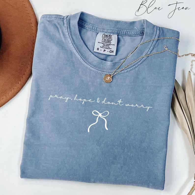 May include: A blue jean-colored t-shirt with the words "pray, hope & don't worry" in white script, along with a bow graphic. A gold necklace with a pendant is draped across the shirt. The shirt is a Comfort Colors brand.
