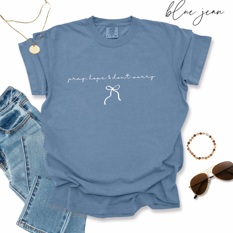 May include: A light blue t-shirt with the words "pray, hope & don't worry" in white script, along with a bow graphic. The words "blue jean" are written in cursive at the top. Accessories include a gold necklace, sunglasses, and a beaded bracelet.