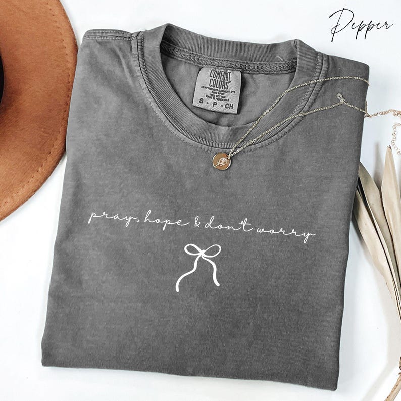 May include: A gray Comfort Colors t-shirt with the text "pray, hope & don't worry" in white script. A silver necklace with a small pendant is draped across the shirt. The shirt is size S-P-CH.