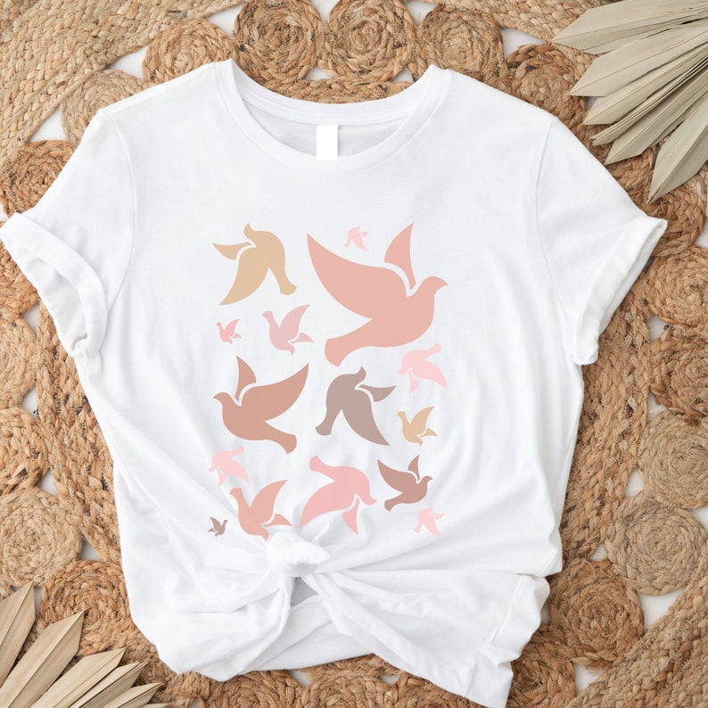 May include: White t-shirt with a design of multiple pink, brown, and beige doves in flight.