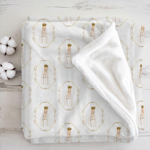 May include: White baby blanket with a repeating pattern of a crowned figure in a gold frame. The blanket has a soft, plush texture and a folded corner. Two cotton buds are visible on the left side.