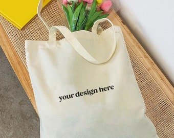 May include: Cream-colored tote bag with the text "your design here" in black. The bag contains a bouquet of pink, white, and yellow tulips. The bag is sitting on a wooden bench.
