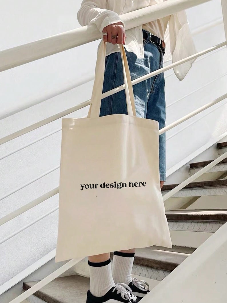 Puede incluir: Bolso tote de color crema con el texto "your design here" en negro. El bolso tiene asas largas. Una persona con vaqueros azules, camisa blanca y zapatillas deportivas blancas y negras sostiene el bolso en una escalera.