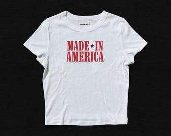 Könnte beinhalten: Weißes T-Shirt mit dem Text "MADE IN AMERICA" in Rot, mit einem blauen Stern. Das T-Shirt hat einen Rundhalsausschnitt und kurze Ärmel. Der Text ist mittig auf der Vorderseite des Shirts platziert.