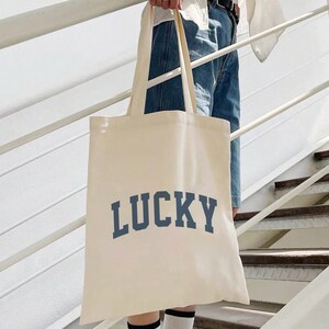 Puede incluir: Bolso tote de color crema con la palabra "LUCKY" impresa en letras de bloque azul marino. El bolso tiene asas largas y se sostiene en una escalera. La persona lleva vaqueros azules, calcetines blancos y zapatillas deportivas blancas y negras.