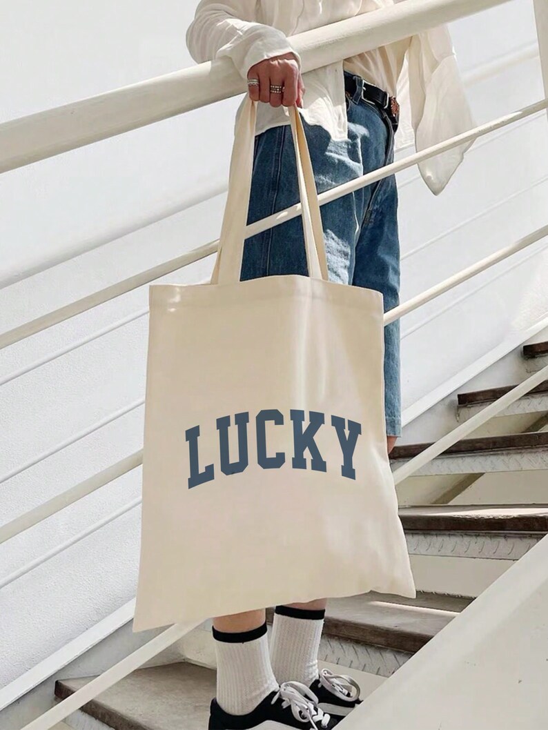 Puede incluir: Bolso tote de color crema con la palabra "LUCKY" impresa en letras de bloque azul marino. El bolso tiene asas largas y se sostiene en una escalera. La persona lleva vaqueros azules, calcetines blancos y zapatillas deportivas blancas y negras.