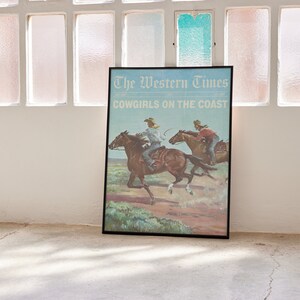 May include: Framed poster titled "The Western Times" with the text "Cowgirls on the Coast." The artwork depicts two cowgirls riding horses across a landscape. The poster is in a black frame and leans against a wall.