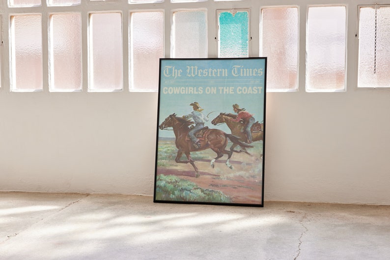 May include: Framed poster titled "The Western Times" with the text "Cowgirls on the Coast." The artwork depicts two cowgirls riding horses across a landscape. The poster is in a black frame and leans against a wall.