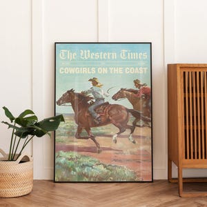 May include: A framed vintage poster titled "The Western Times" with the headline "Cowgirls on the Coast." The artwork depicts two cowgirls riding horses across a landscape. The poster is framed in black and displayed against a white wall.