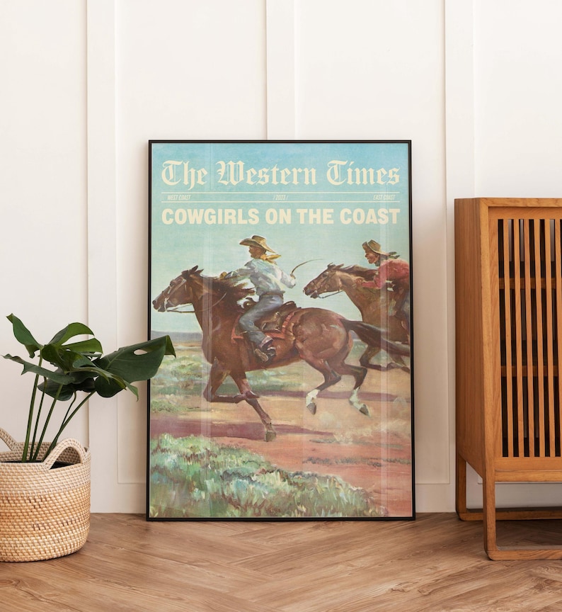 May include: A framed vintage poster titled "The Western Times" with the headline "Cowgirls on the Coast." The artwork depicts two cowgirls riding horses across a landscape. The poster is framed in black and displayed against a white wall.