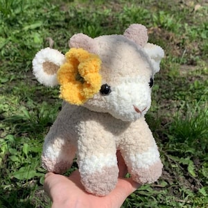Op de afbeelding: Een handgemaakte gehaakte koeienknuffel in cr&egrave;me, wit en bruin. De koe heeft een gele bloemaccent en zwarte veiligheidsoogjes. De knuffel is zacht en pluizig, ideaal voor een kinderspeelgoed of kinderkamerdecoratie. De tekst "fiya.crochet" is zichtbaar.