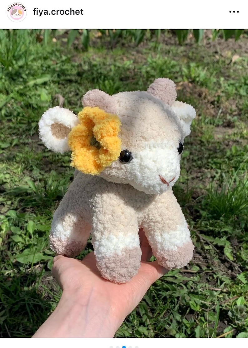 Op de afbeelding: Een handgemaakte gehaakte koeienknuffel in cr&egrave;me, wit en bruin. De koe heeft een gele bloemaccent en zwarte veiligheidsoogjes. De knuffel is zacht en pluizig, ideaal voor een kinderspeelgoed of kinderkamerdecoratie. De tekst "fiya.crochet" is zichtbaar.