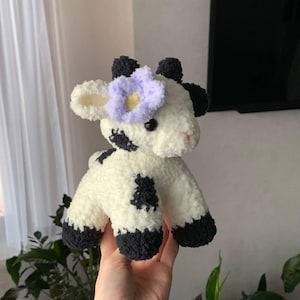 Op de afbeelding: Een handgemaakte pluche koe, wit met zwarte vlekken en hoeven. De koe heeft zwarte oren en een lichtpaarse bloem op het hoofd. Dit zachte speelgoed is ideaal voor kinderen en is gemaakt van zacht materiaal.