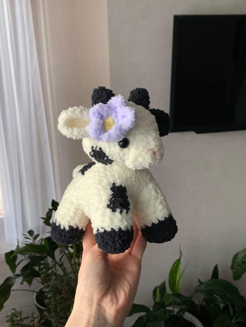 Op de afbeelding: Een handgemaakte pluche koe, wit met zwarte vlekken en hoeven. De koe heeft zwarte oren en een lichtpaarse bloem op het hoofd. Dit zachte speelgoed is ideaal voor kinderen en is gemaakt van zacht materiaal.