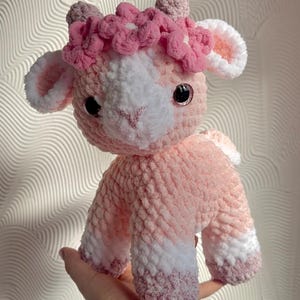 Op de afbeelding: Een handgemaakte gehaakte lammetjespop in roze en wit. Het lam heeft een bloemenkrans en zwarte veiligheidsoogjes. De pop is zacht en pluche, perfect als kinderspeelgoed of voor de kinderkamerdecoratie.
