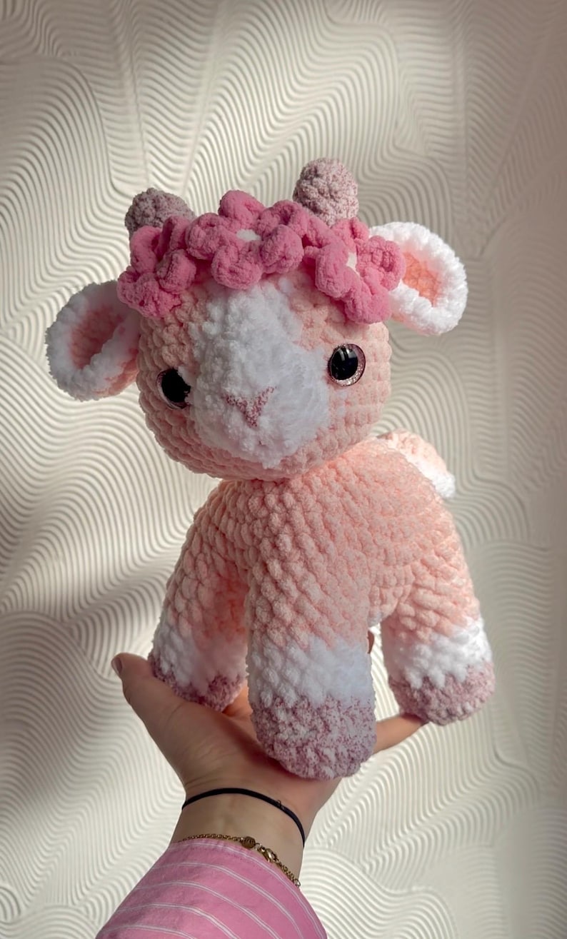 Op de afbeelding: Een handgemaakte gehaakte lammetjespop in roze en wit. Het lam heeft een bloemenkrans en zwarte veiligheidsoogjes. De pop is zacht en pluche, perfect als kinderspeelgoed of voor de kinderkamerdecoratie.
